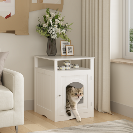 A white wooden cat litter box enclosure used as a side table next to a sofa, with a tabby cat stepping out of the arched door opening in a bright living room.