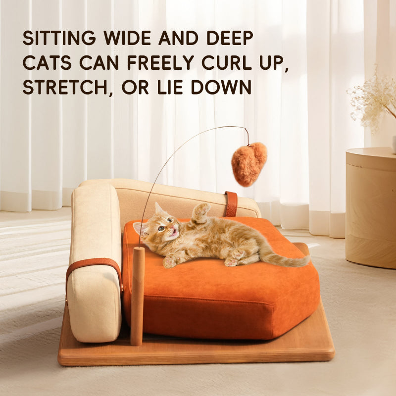 An playful orange kitten lying on a wide orange pet couch bed, reaching up to play with an attached hanging plush toy in a bright room.