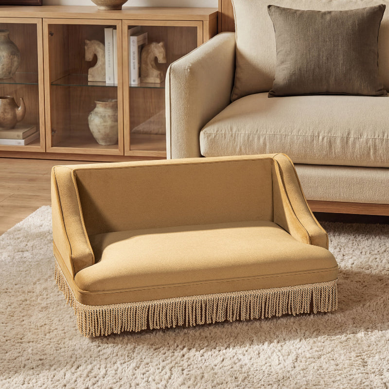 A front view of a retro mustard yellow velvet cat sofa with tassel fringe trim sitting on a textured rug in a cozy living room.