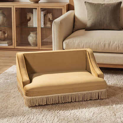 A front view of a retro mustard yellow velvet cat sofa with tassel fringe trim sitting on a textured rug in a cozy living room.