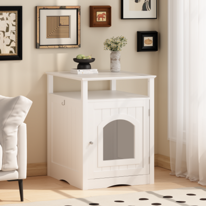 A white wooden cat litter box enclosure styled as a side table in a cozy room corner, topped with decorative items and an open shelf.