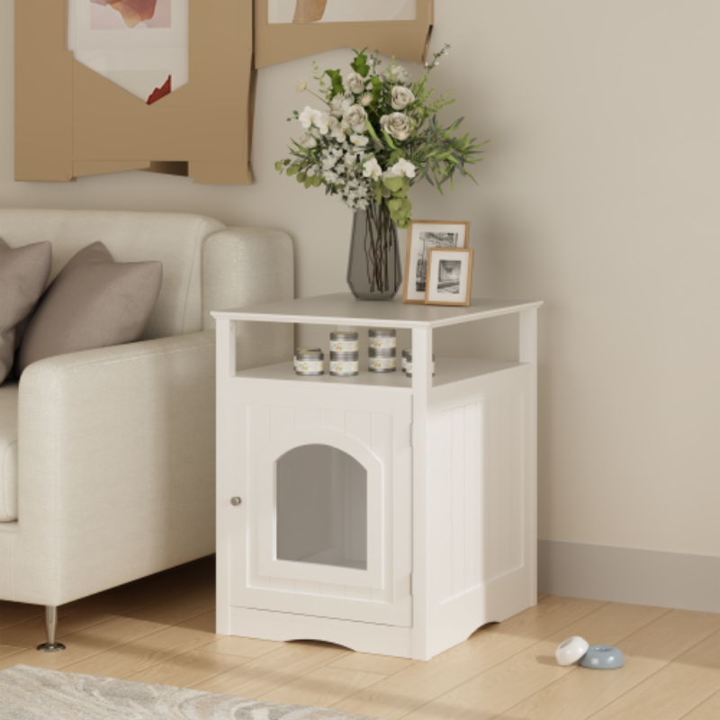 A white wooden cat litter box enclosure placed beside a sofa as a side table, with flowers and decor on top and an open storage shelf.